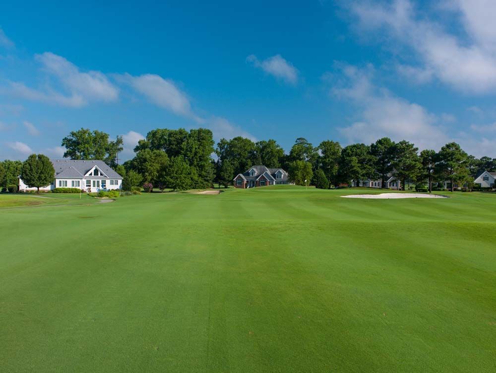 A green golf course with houses in the background under a blue sky. The houses are mostly white and gray.