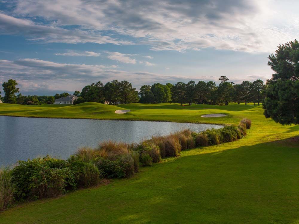 Lush green golf course with water hazard, bushes, and trees under a cloudy blue sky.