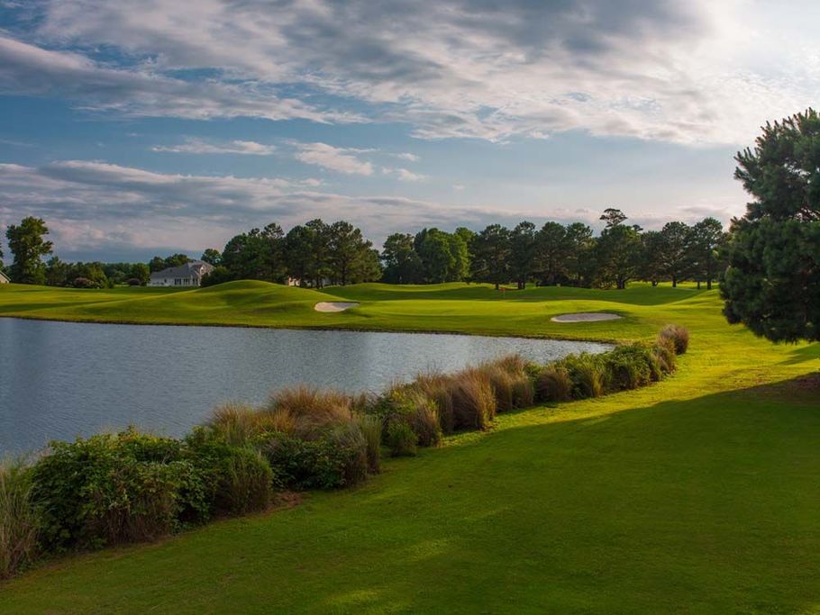 Lush green golf course with water hazard, bushes, and trees under a cloudy blue sky.