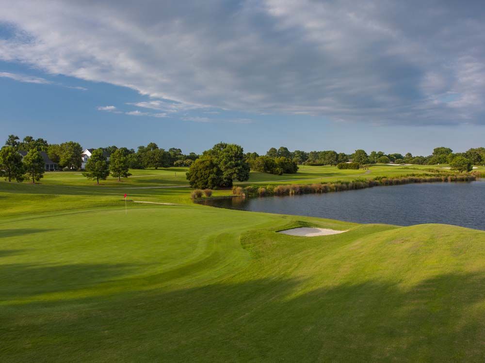Green golf course with a lake, sand trap, and cloudy sky.