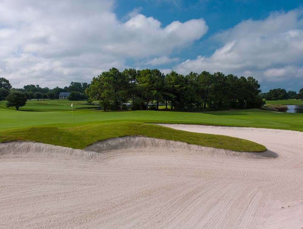A golf course scene with a sand trap in the foreground, green grass, and trees under a cloudy sky.