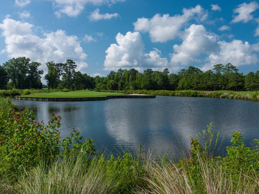 A scenic golf course with a reflective lake under a blue sky filled with fluffy white clouds. Green foliage frames the water.