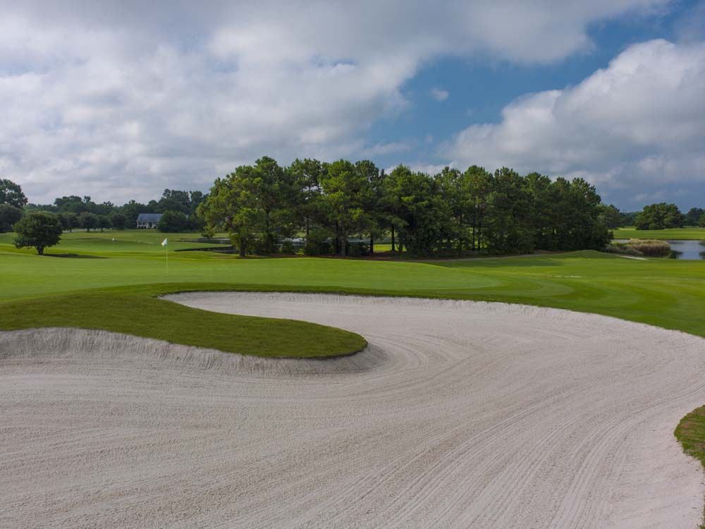 A golf course with a sand trap in the foreground, lush green fairways, and trees under a partly cloudy sky.