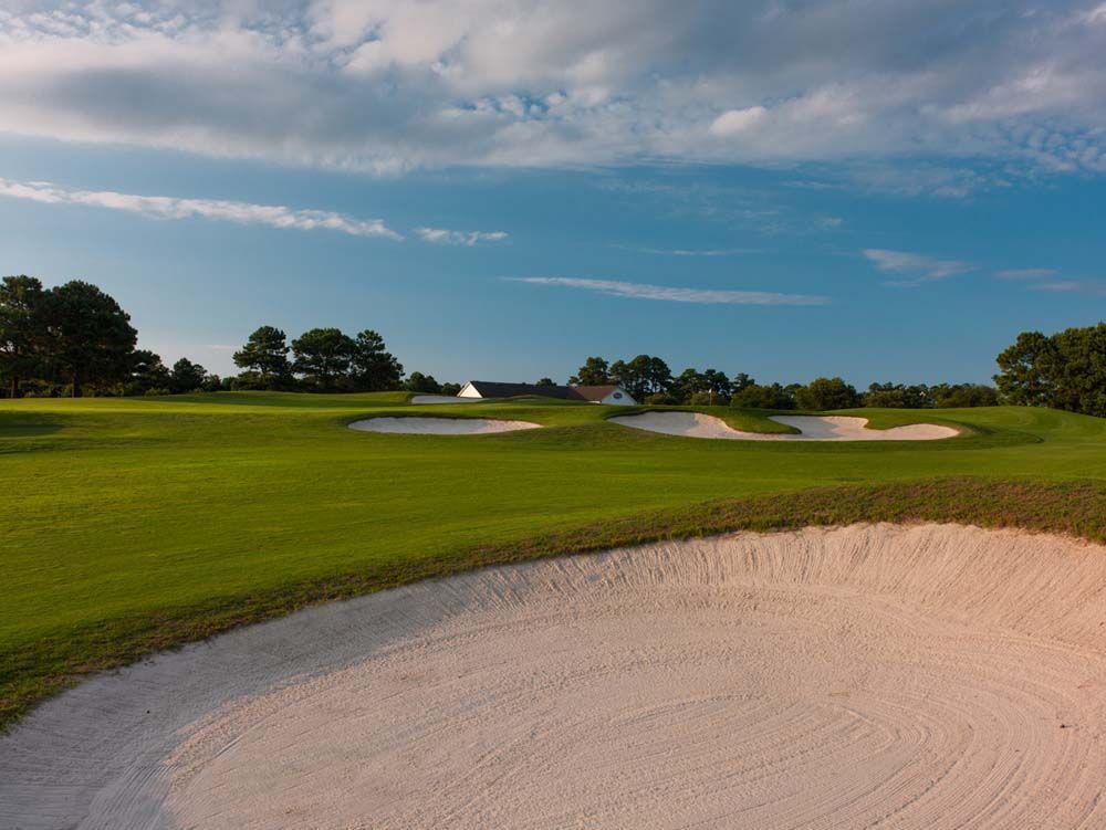 A golf course scene with a sand trap in the foreground, green grass, and bunkers in the background under a blue sky with clouds.