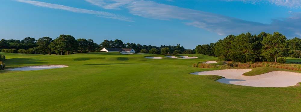 A green golf course under a clear blue sky. White sand bunkers are visible with trees in the background.