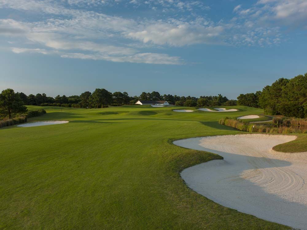A golf course with green grass, sand traps, and a few trees. Blue sky with clouds visible in the background.