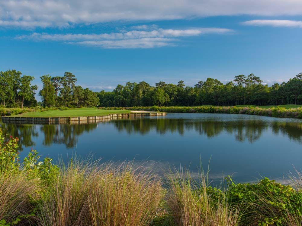 A tranquil lake on a golf course reflecting trees and a blue sky. Tall grasses frame the foreground.
