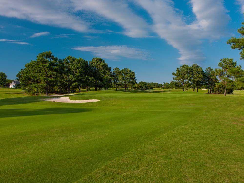 Green golf course with sand trap, trees, and blue sky with clouds.