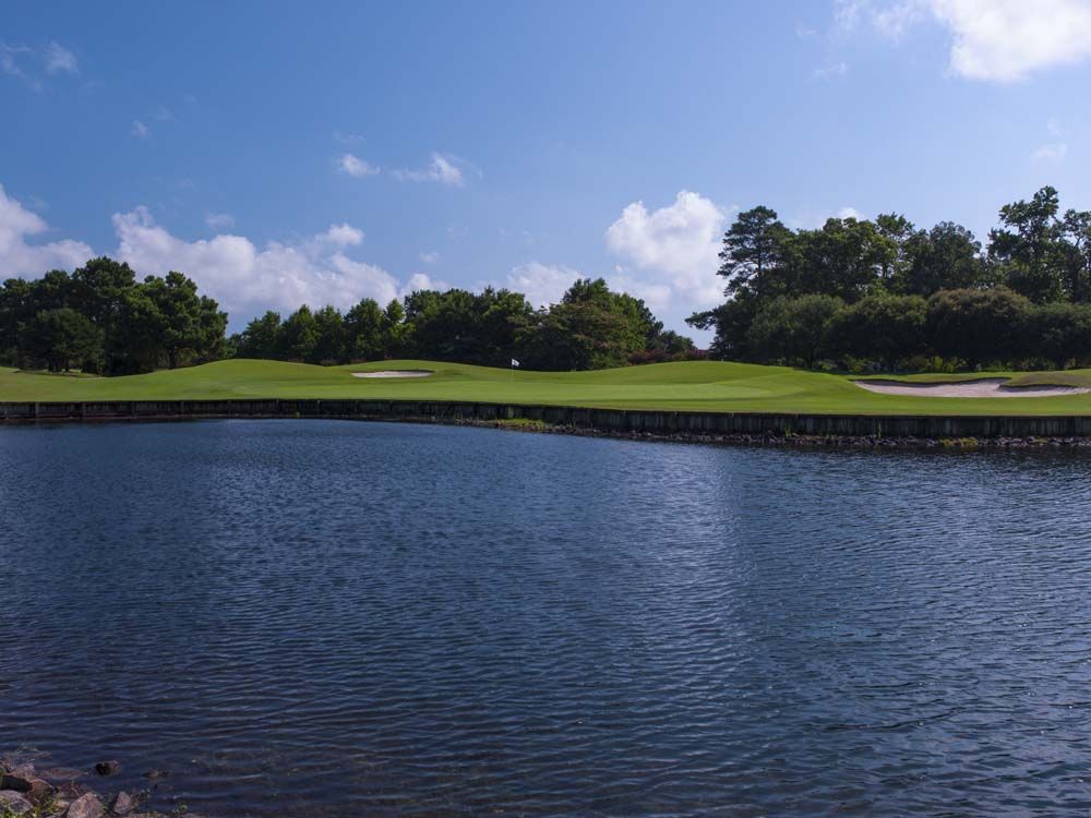 A golf course with a green and water feature under a blue sky. The green has a flag in the center.