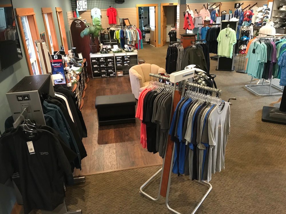 Interior view of a clothing store with racks of shirts, jackets, and accessories for sale. Includes wooden floors and a seating area.