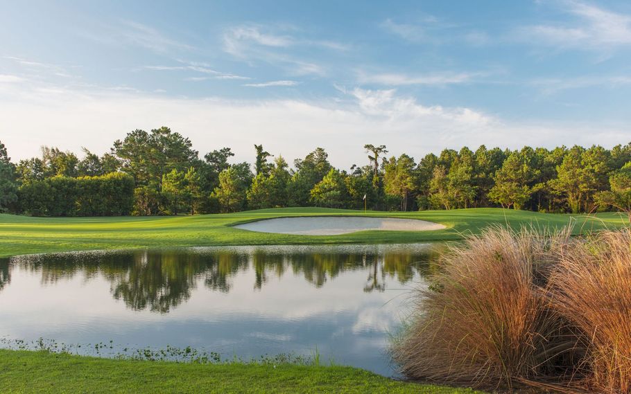 Golf course scene with water hazard reflecting trees and blue sky. Green grass surrounds the water and a sand trap.