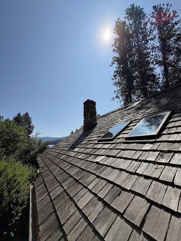 Old, weathered wooden shingle roof with chimney, skylights, and a bright sun in a blue sky.