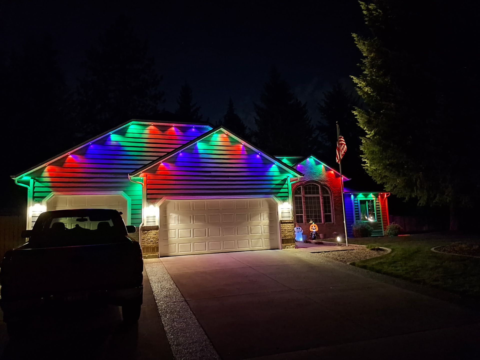 House decorated with red, green, and blue Christmas lights on roof and around the door at night. A car is parked in the driveway.