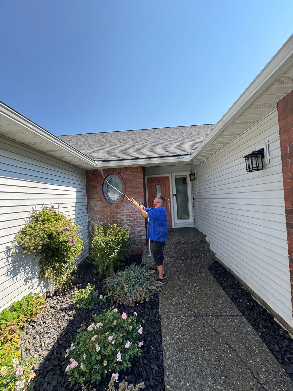 Man spraying water on the roof of a house. Blue shirt, black shorts, sunny day.