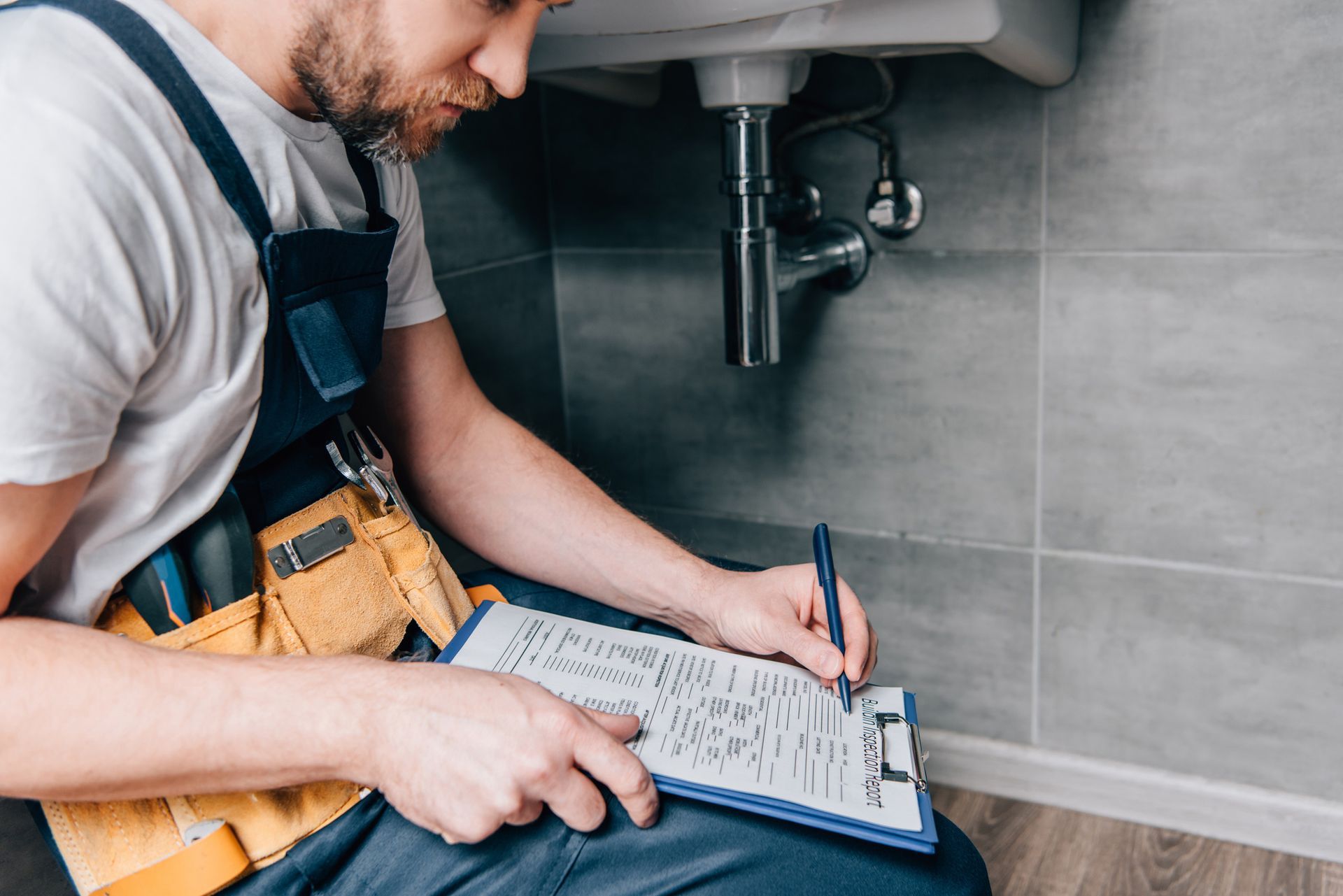 A plumber is sitting under a sink writing on a clipboard.
