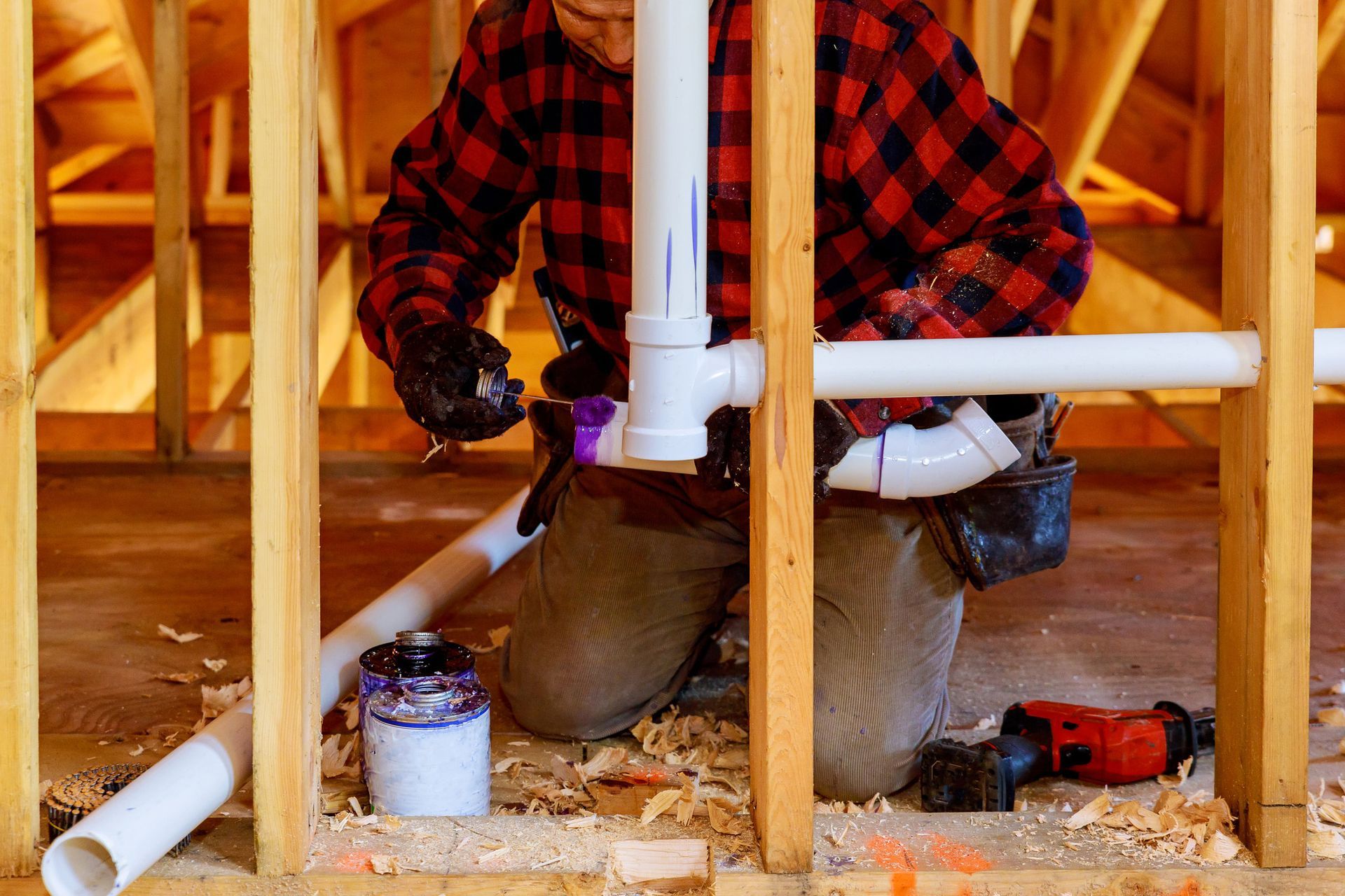 A man in a plaid shirt is working on pipes in a house under construction.