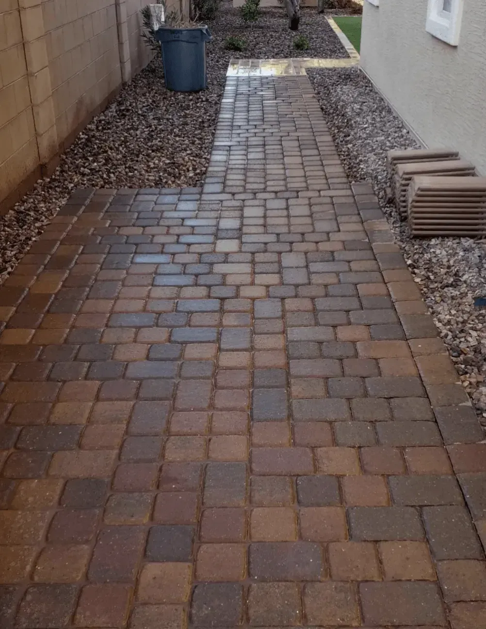 A brick walkway leading to a house with a trash can in the background