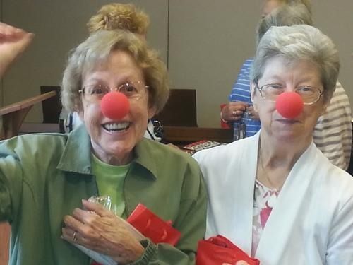 Two women wearing red clown noses, smiling in a room. One waves while holding a gift.