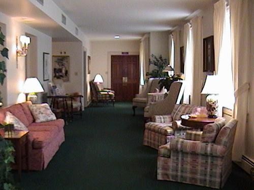 Lounge area with pink couch, plaid chairs, lamps, and windows with white curtains, on a green carpet.