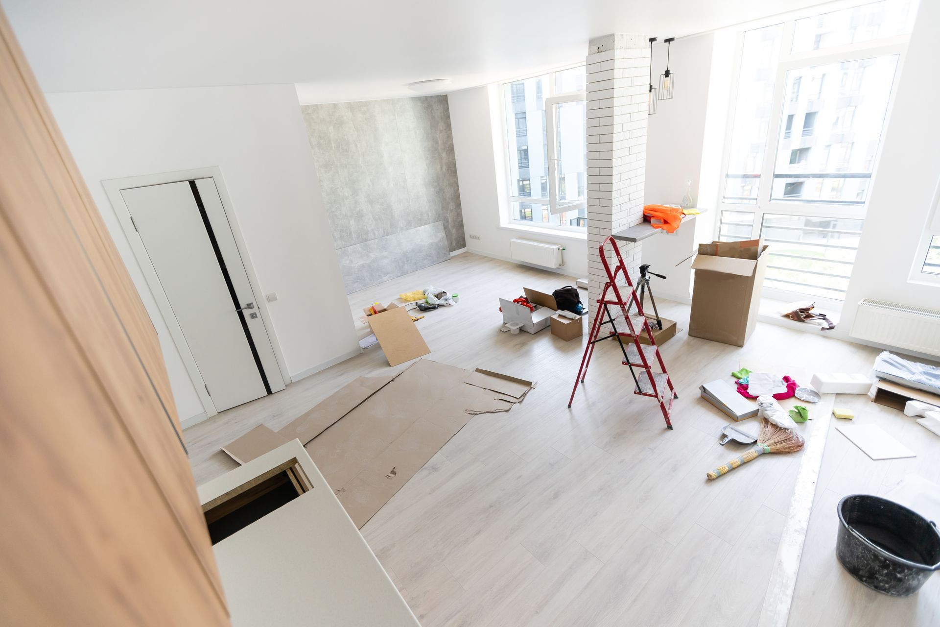 Bright renovation room with ladder, tools, and scattered materials on light wood floor.