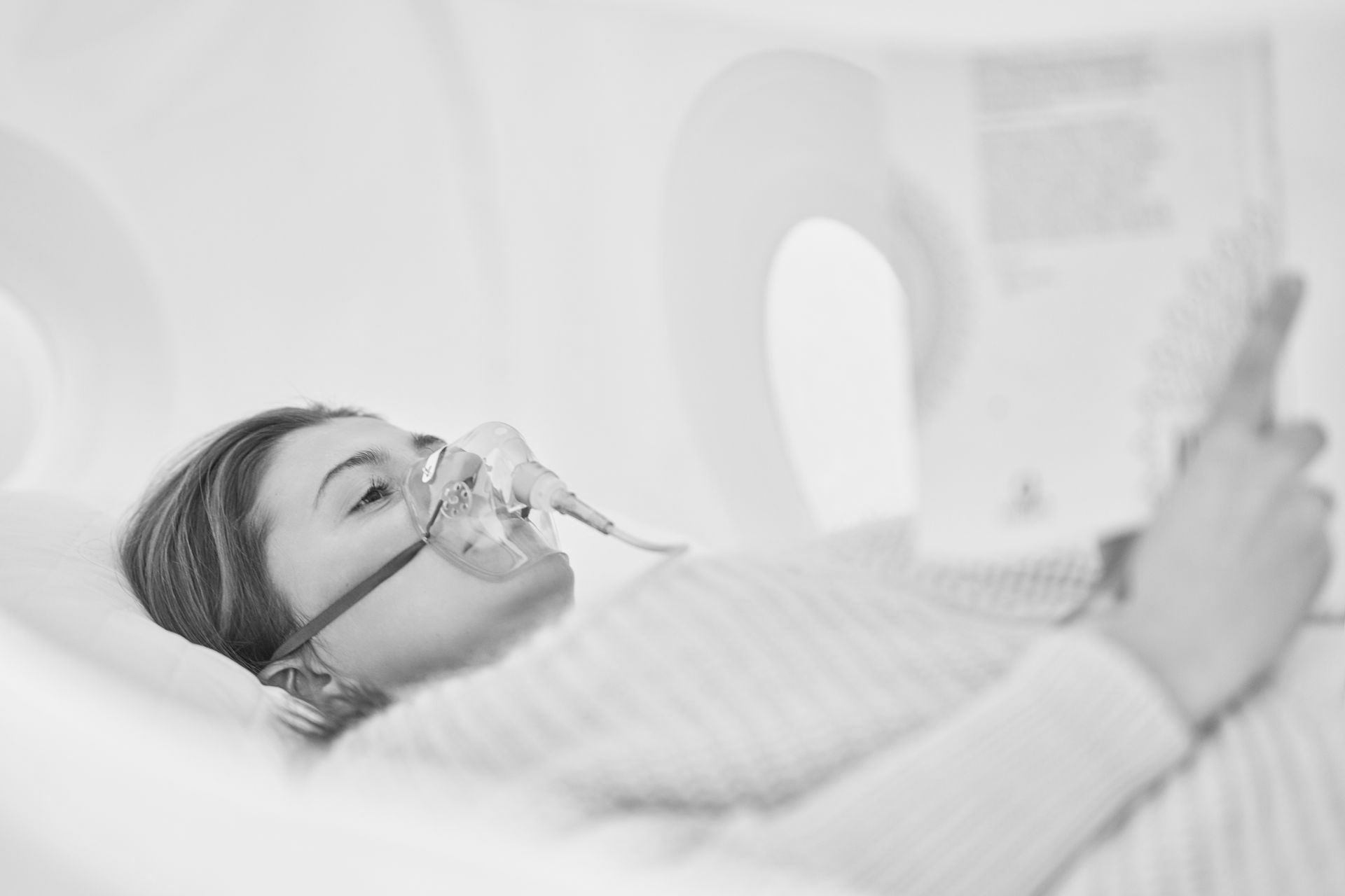 Woman with oxygen mask lying down, possibly in a medical setting, reading a document.