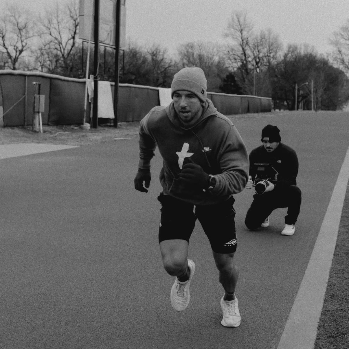 Runner in hoodie, hat, and shorts, sprinting on asphalt with coach squatting behind.