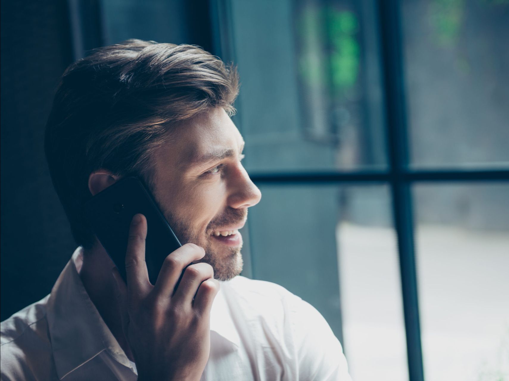 A man is sitting in front of a window talking on a cell phone.