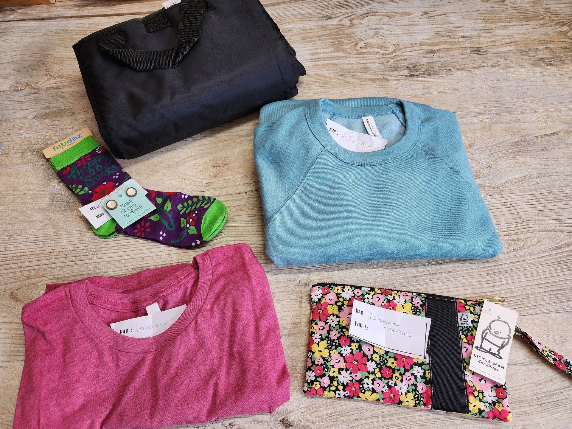Black tote bag, teal sweatshirt, pink shirt, floral wallet, and socks with tags on a wooden surface.