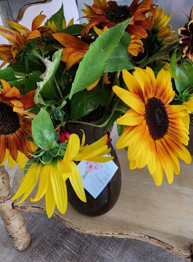 A bouquet of yellow and orange sunflowers in a brown vase with a tag, set on wood.