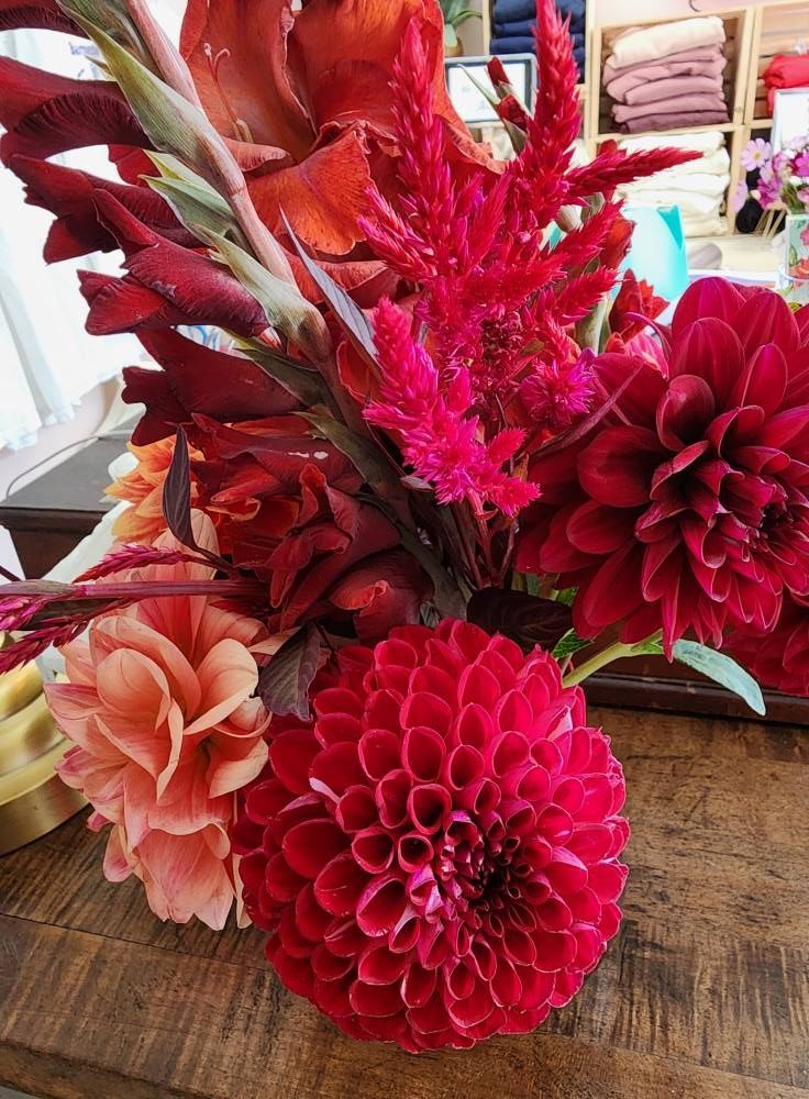 Vibrant red and orange flower arrangement on a wooden table. Dahlias, gladiolus, and celosia are prominent.