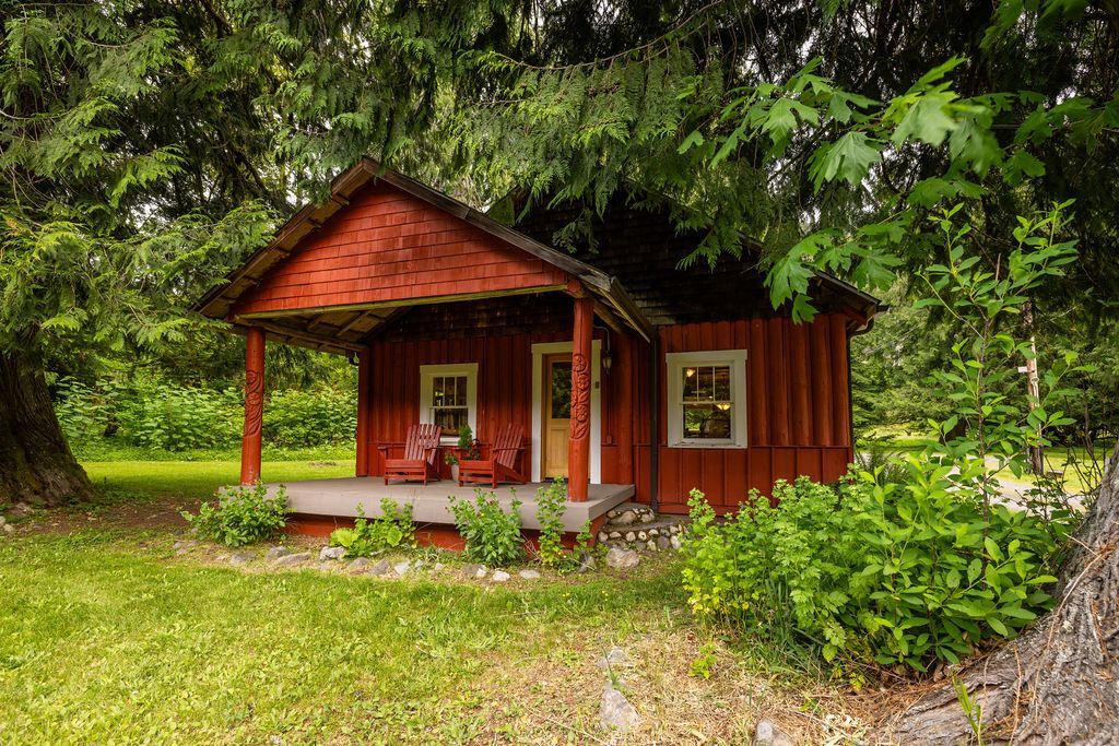 Red wooden cabin nestled under green trees, with porch and two chairs. Lush grass surrounds it.