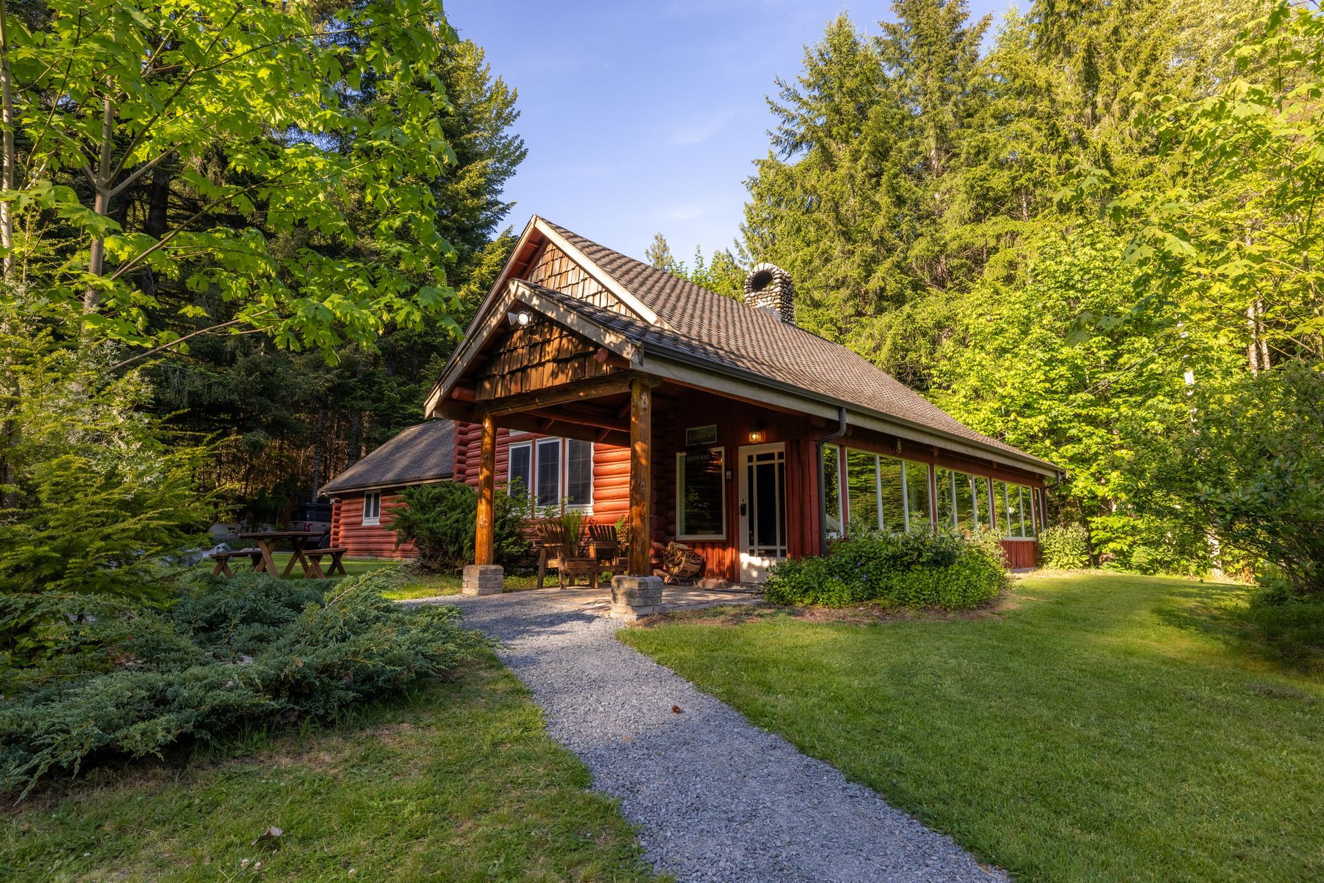 A rustic, red cabin with a stone path leading to the entrance, surrounded by lush green trees and grass on a sunny day.