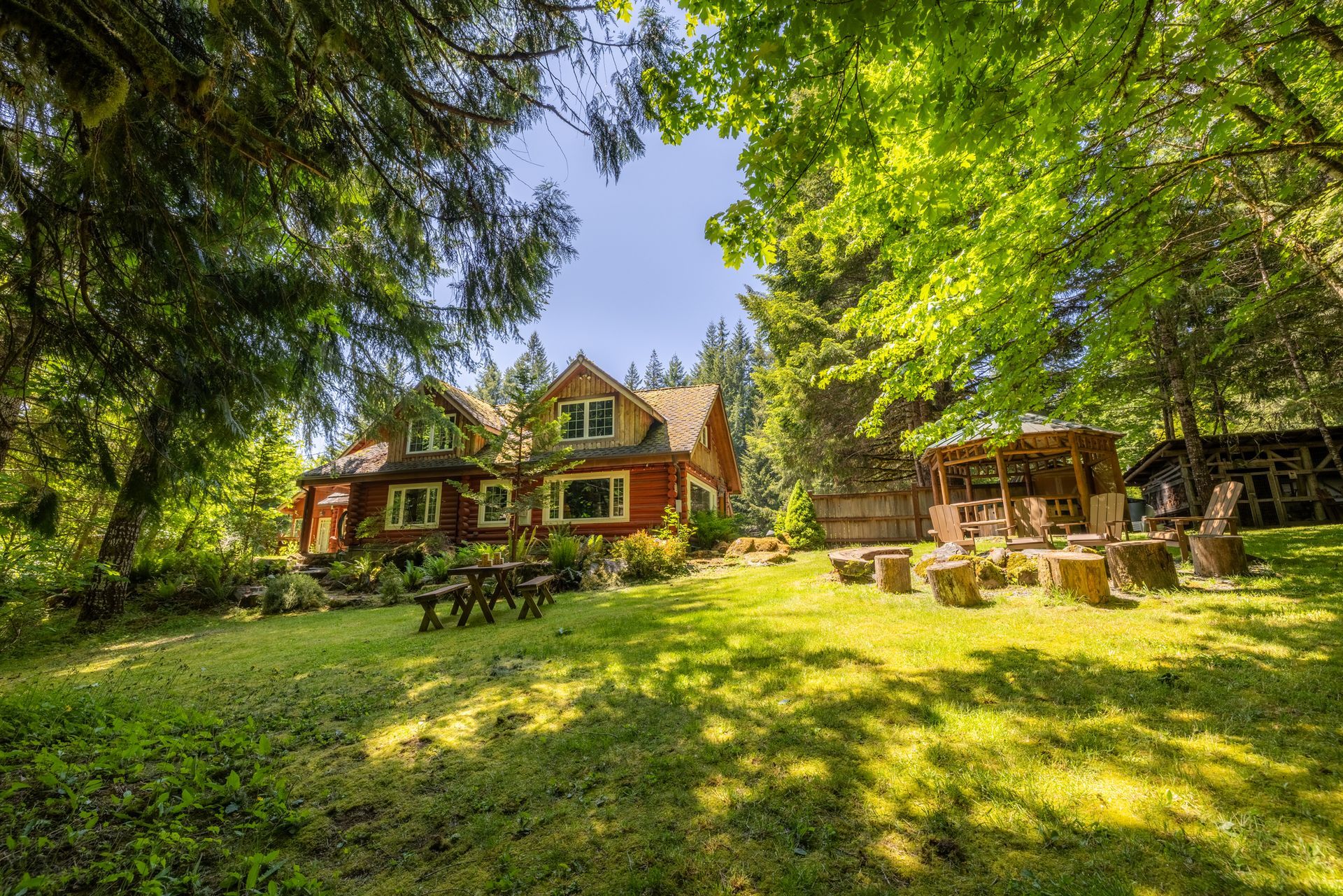Red house with a green yard surrounded by trees on a sunny day. Wooden picnic tables and stone seating in the foreground.