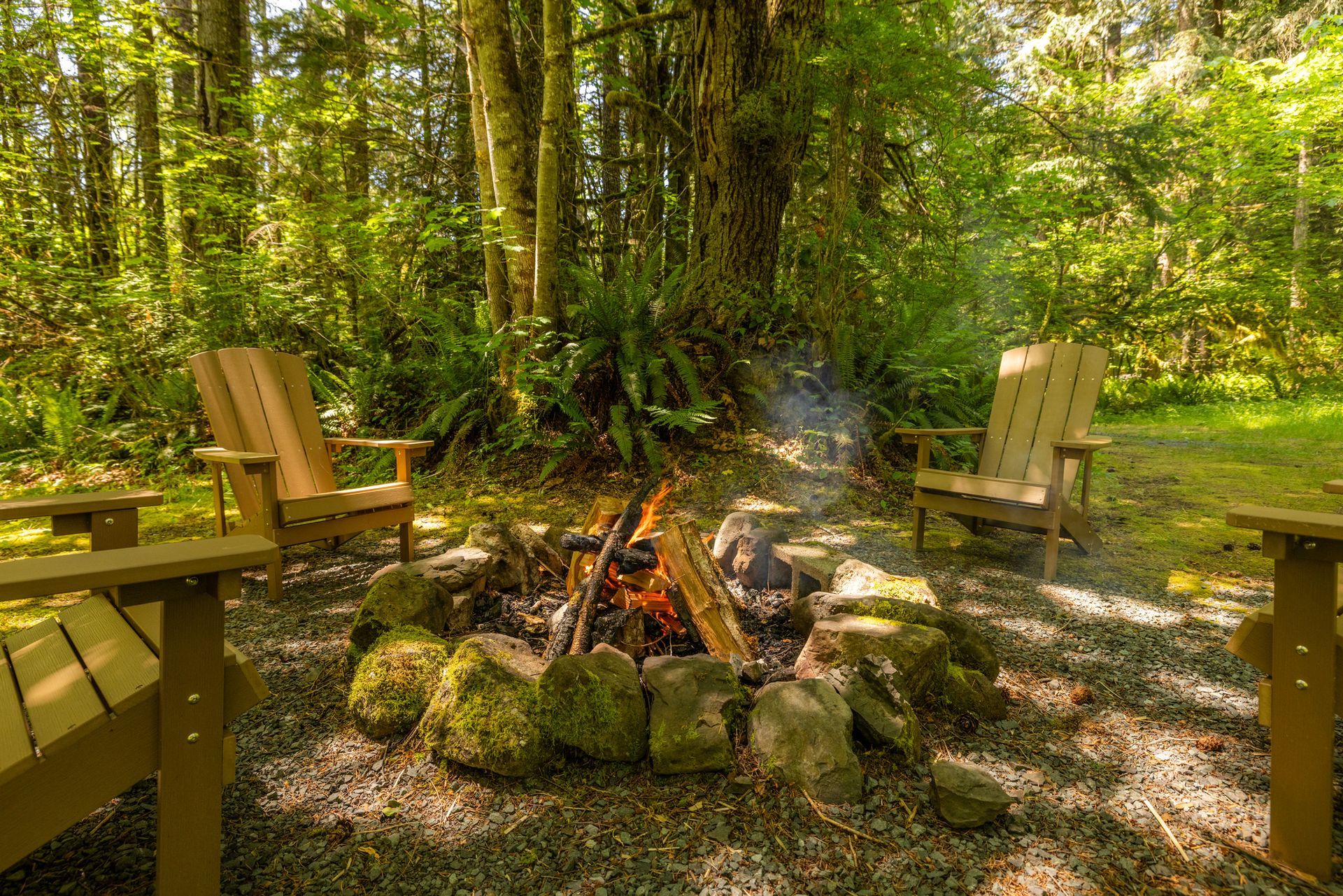 Three wooden chairs around a campfire pit in a forest, smoke rising from the fire.