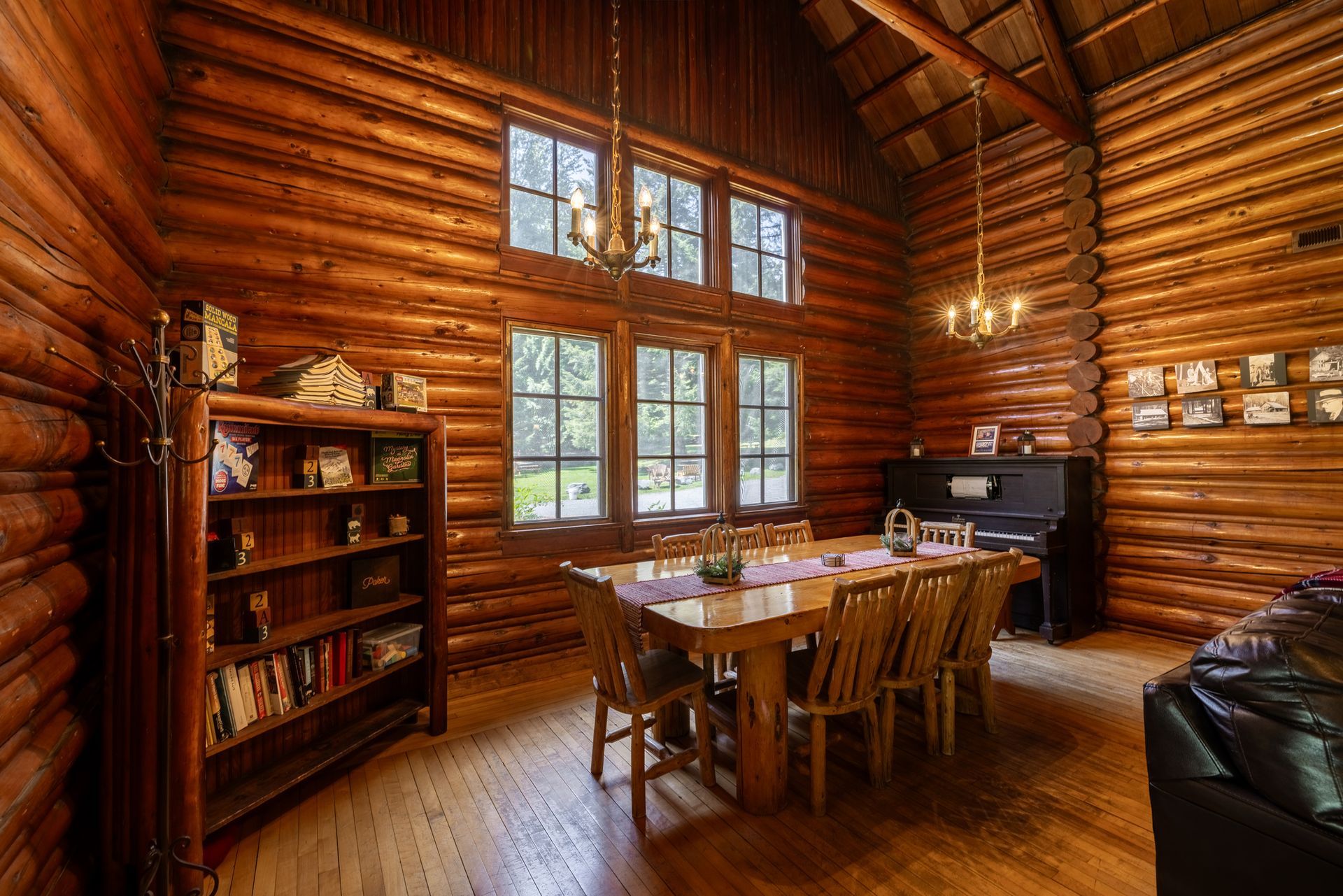 Interior of a log cabin dining room with a large wooden table and chairs, a piano, and a bookshelf. Wooden walls and floors.