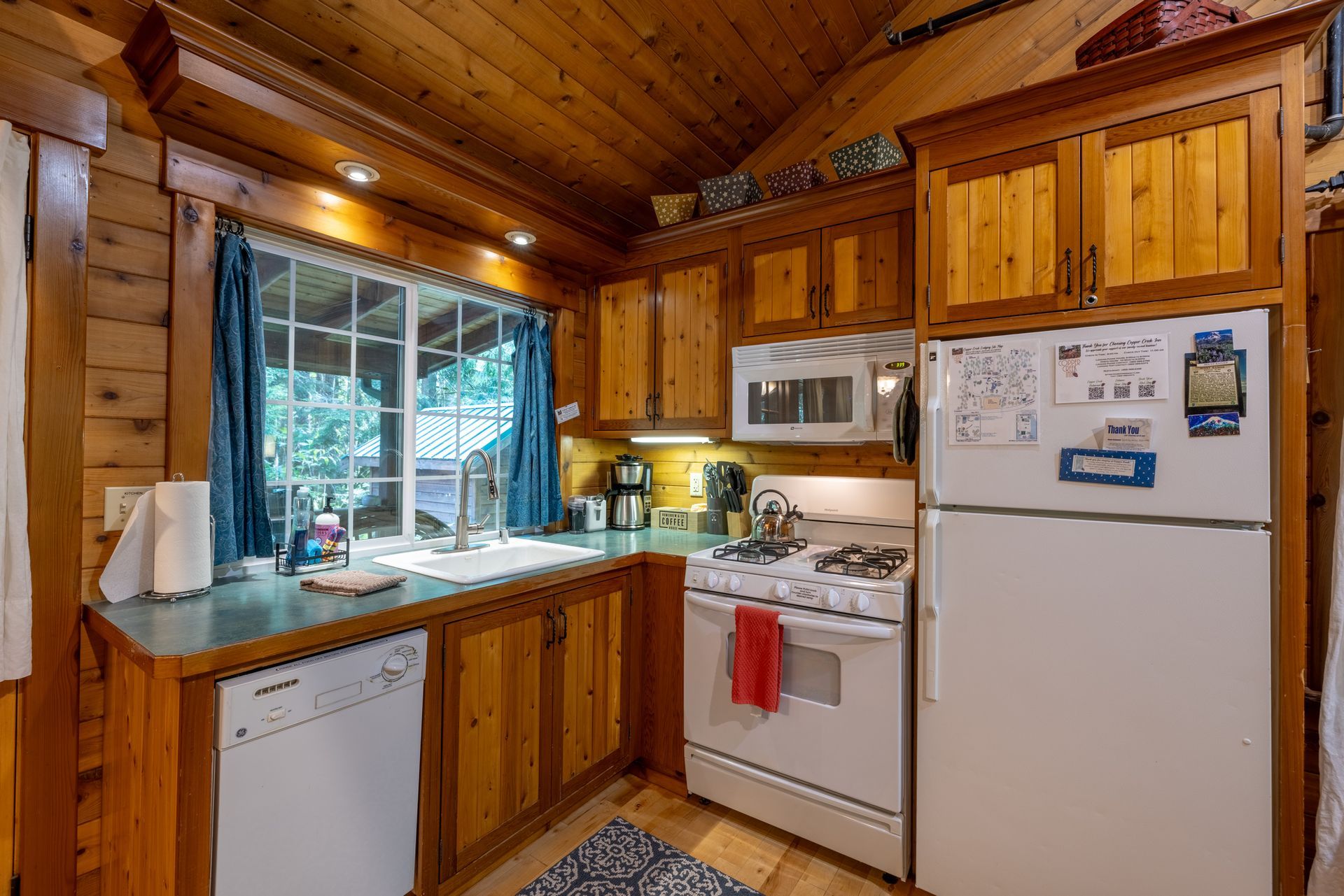 Cozy, rustic kitchen with wooden cabinets, a white stove and refrigerator, and a window overlooking greenery.