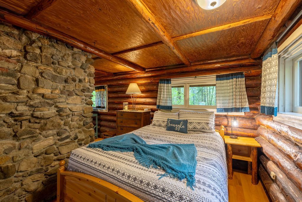 Cozy log cabin bedroom with a bed, stone wall, and wooden furniture. Features natural wood tones, a blue throw, and patterned bedding.