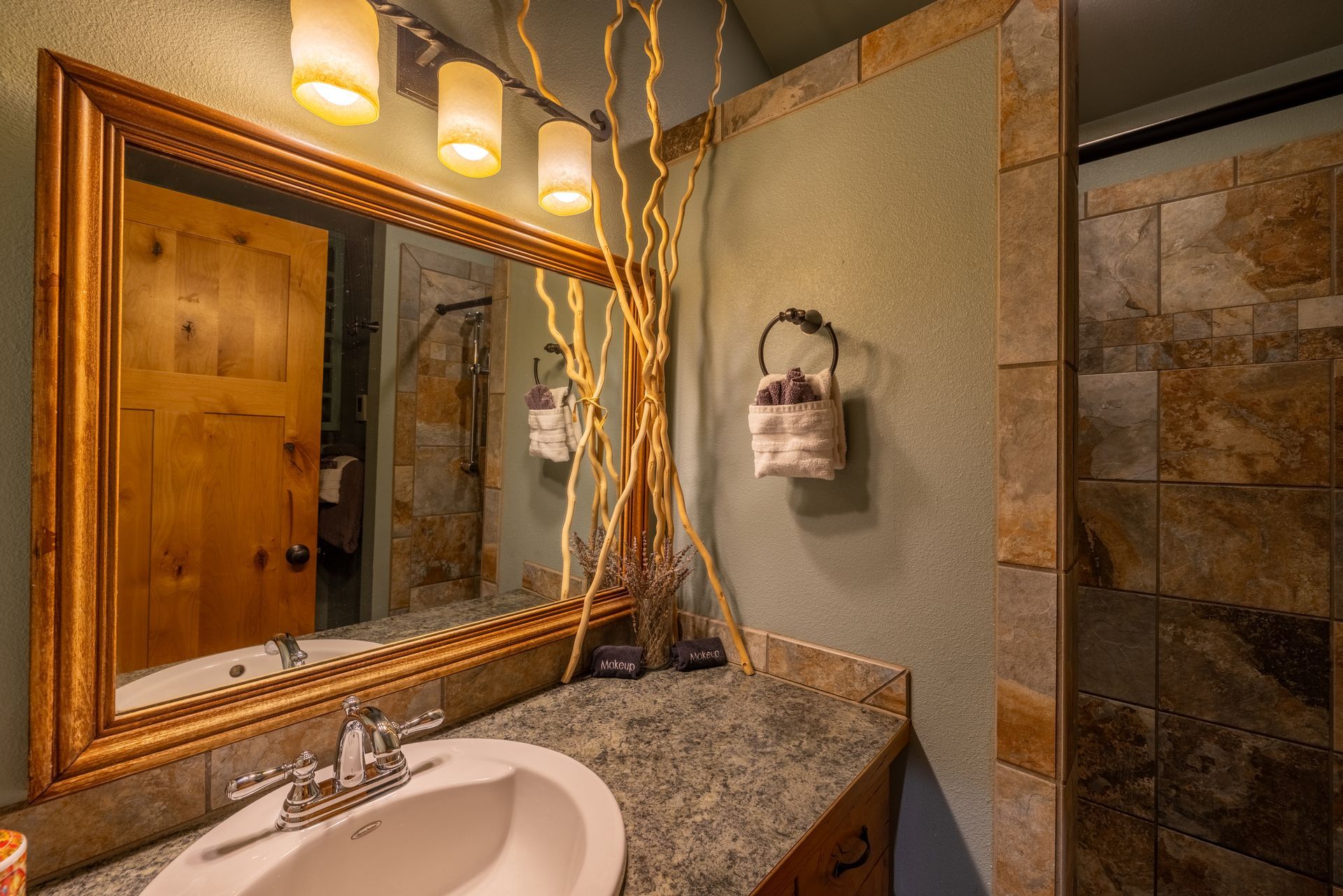 Bathroom with a stone shower and vanity, wooden door reflection in mirror, and decorative branches.
