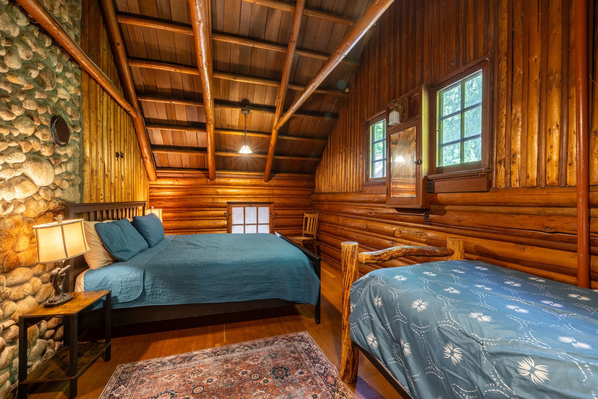 Bedroom in a rustic cabin with wood paneling, stone wall, two beds with blue bedding, and small windows.
