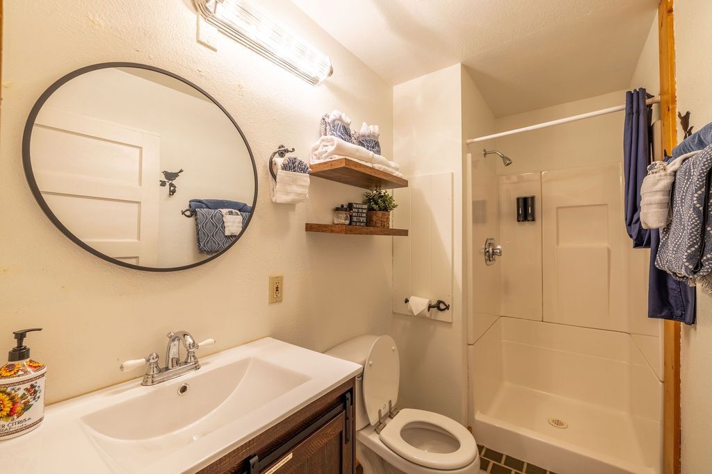 Bathroom with a white sink, toilet, and shower. A round mirror hangs above the sink, with wooden shelves holding towels.