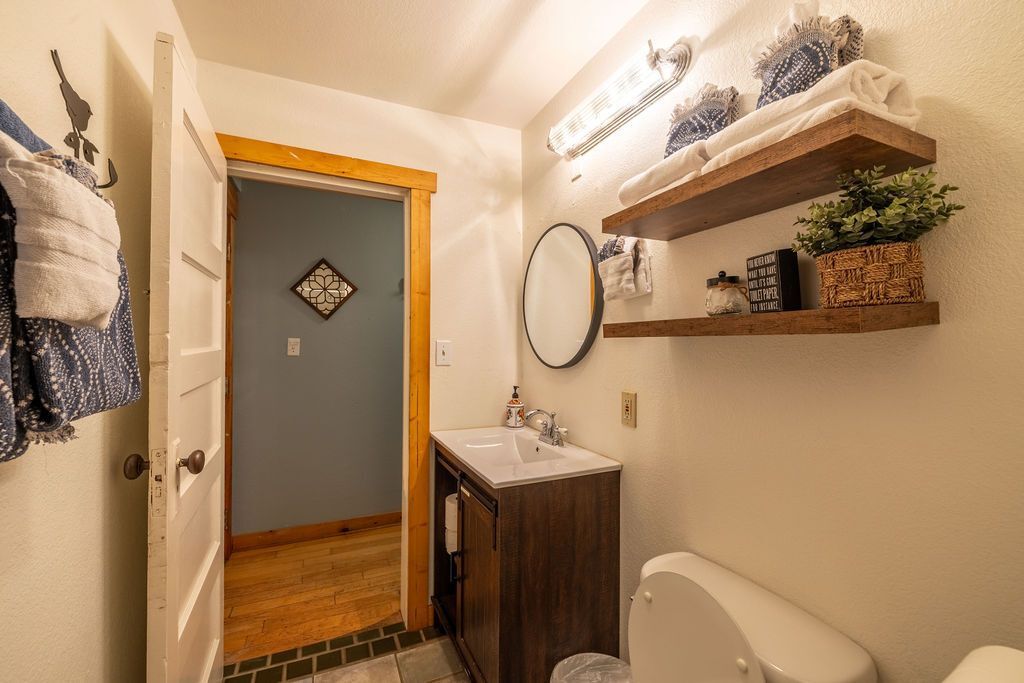 Small bathroom with an open doorway; features a sink, toilet, and shelving with towels and decor.