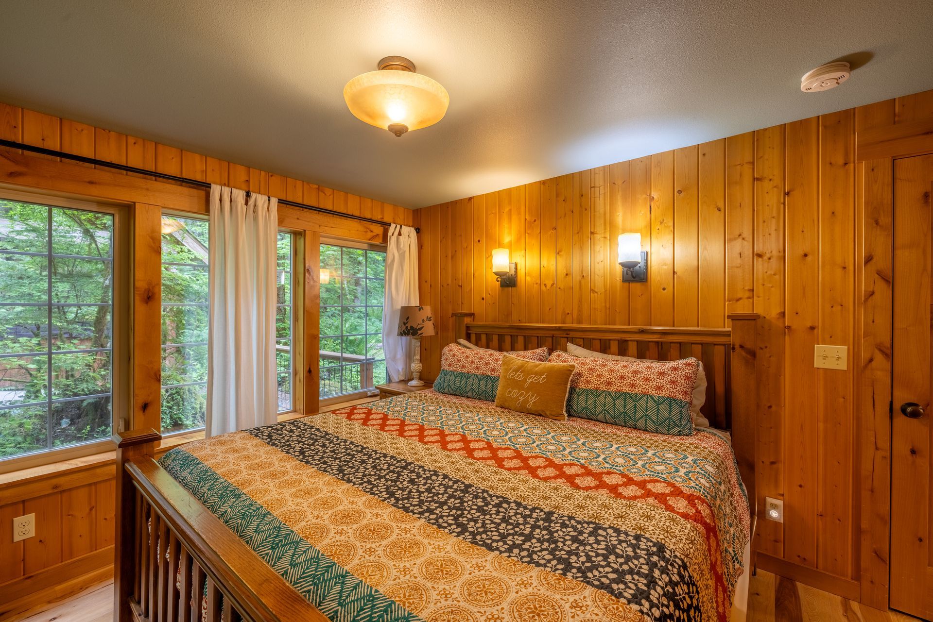 Bedroom with a wooden bed, walls, and trim, windows with white curtains, and a colorful quilt.