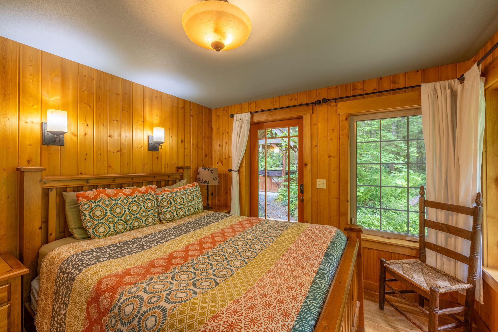 Bedroom with wood-paneled walls, a bed with a colorful quilt, a door to a patio, and a window with a view of trees.