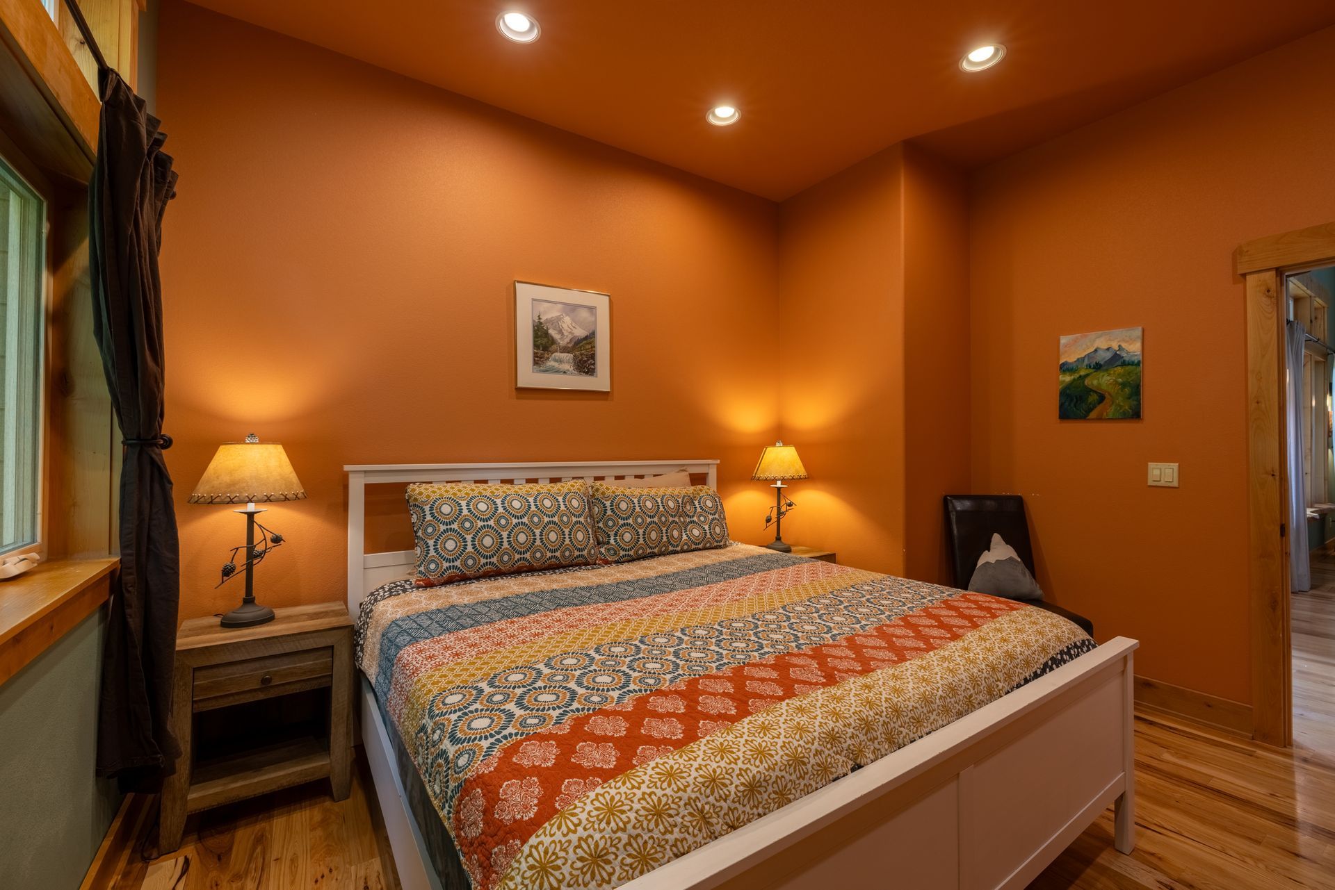 Bedroom with orange walls, white bed frame, patterned bedding, and two bedside lamps on wooden nightstands. A small piece of art hangs on each wall.