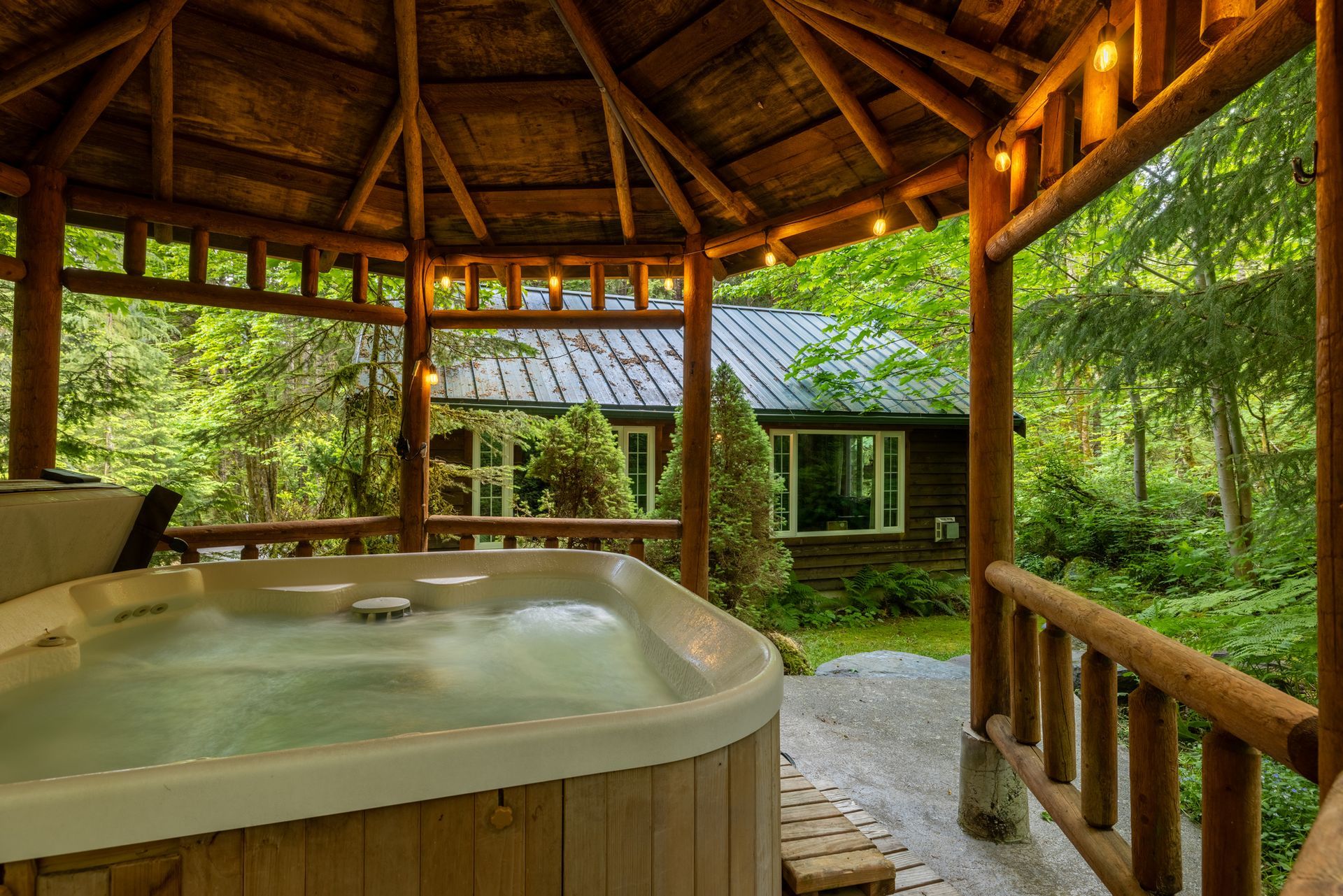 Hot tub on a wooden deck, under a gazebo, overlooking a cabin in a forest. Warm lights and lush green trees surround.