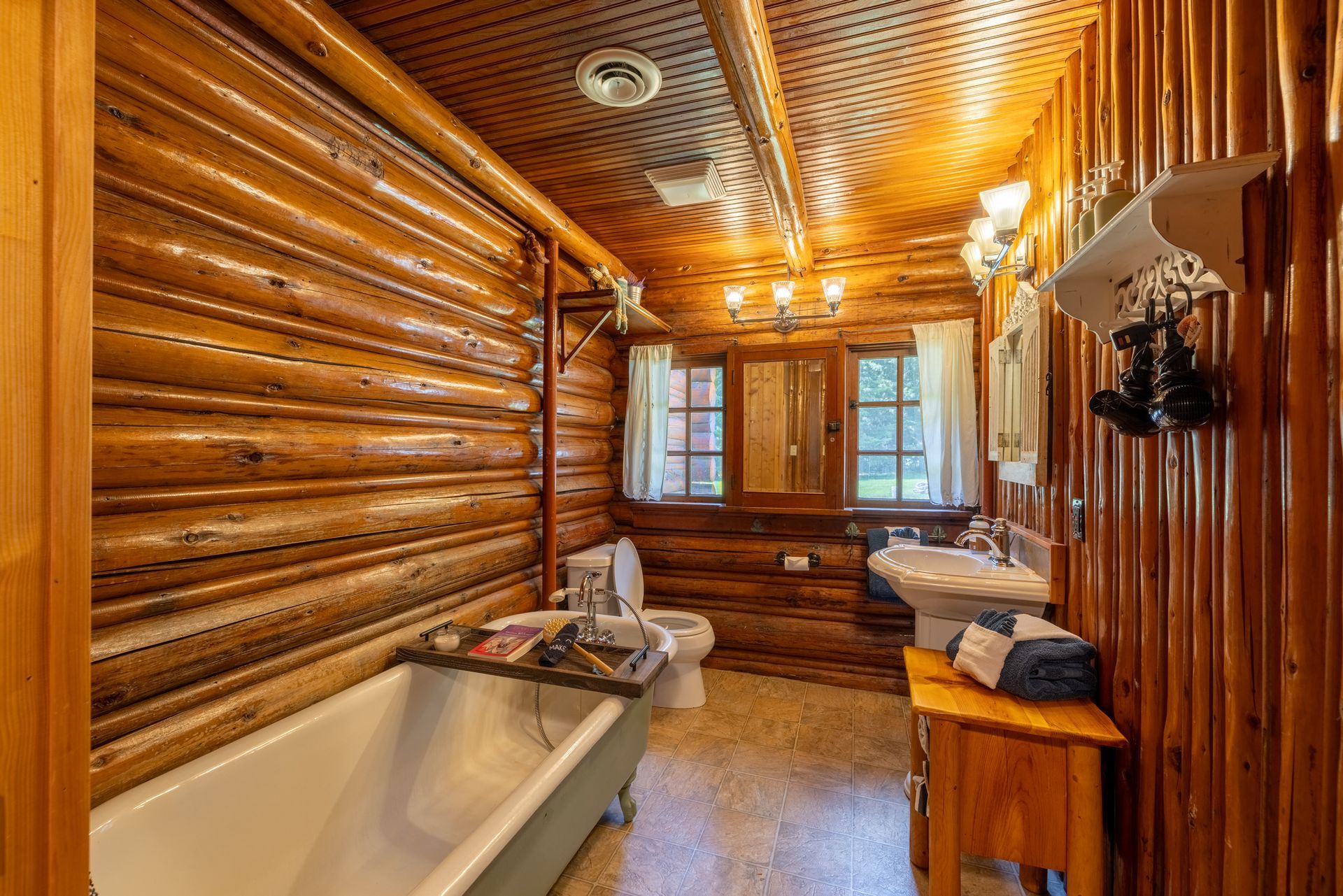 Log-walled bathroom with a claw-foot tub, toilet, sink, and window. Warm wood tones dominate the space, creating a rustic atmosphere.