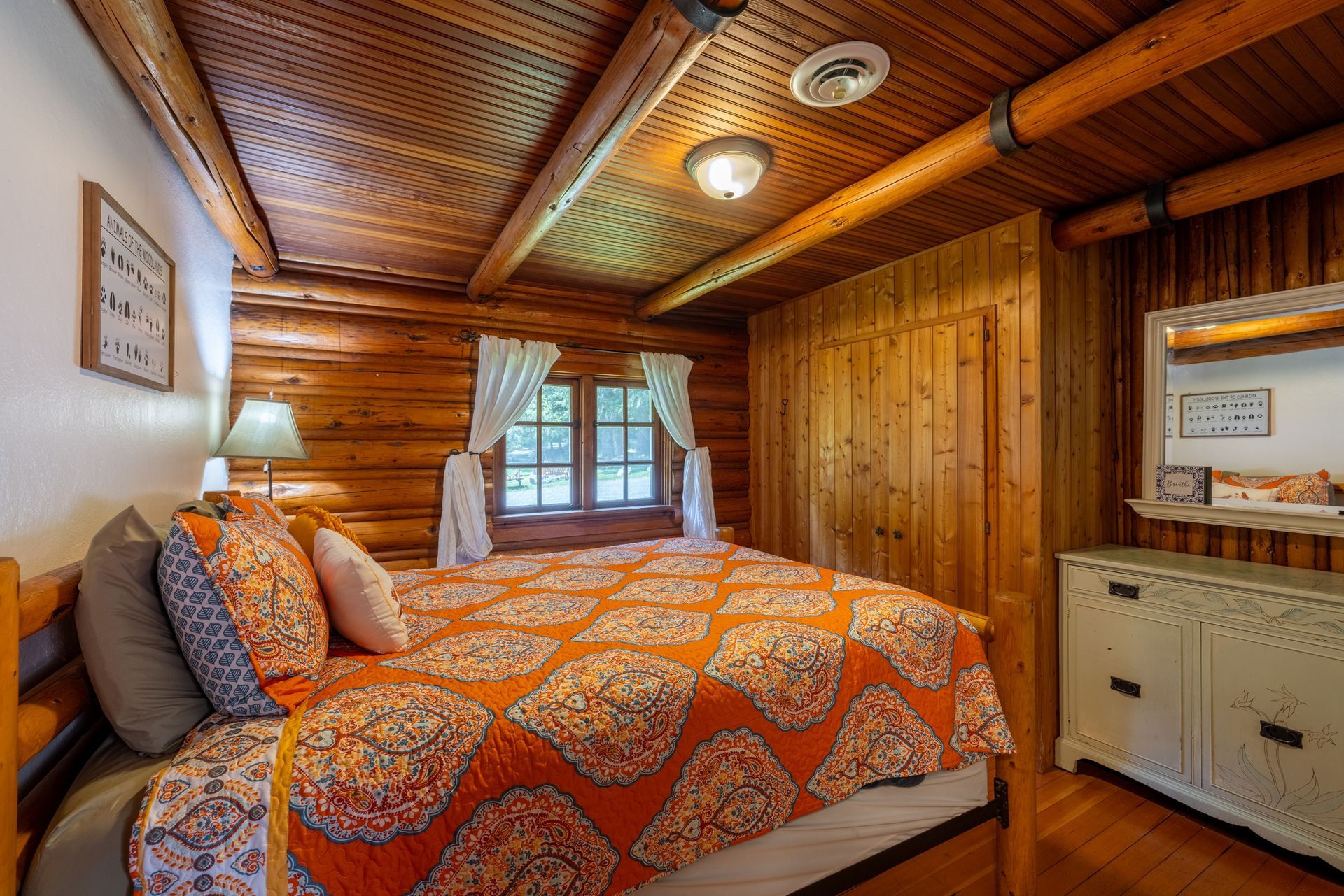 Bedroom with log walls and ceiling.  Bed with orange quilt.  Window with curtains, dresser, and lamp.