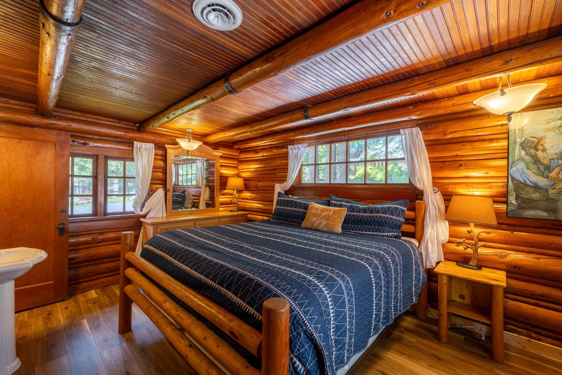 Cozy log cabin bedroom with a bed, windows, and warm wood tones. A bedside table and artwork complete the scene.