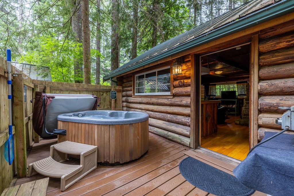 A wooden hot tub sits on a deck beside a log cabin. The cabin door is open, and trees surround the area.