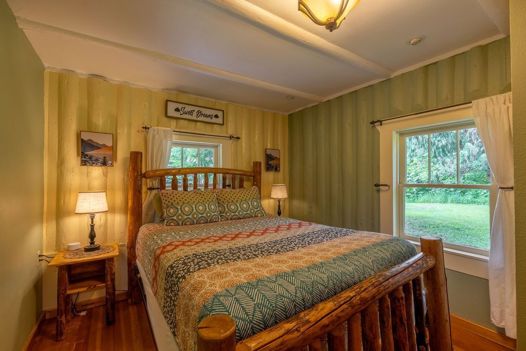 Bedroom with a wooden log bed, bedside table, and two windows overlooking greenery. Yellow and green walls.